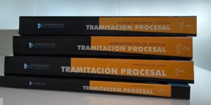 Pila horizontal de los cuatro volúmenes del temario en papel de Tramitación Procesal de Academia ADJ, sobre una superficie reflectante y con el logo de la academia en la pared del fondo